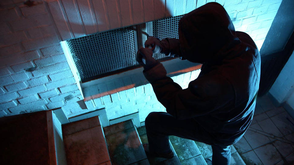 Dunkle Umgebung. Ein Einbrecher versucht ein Kellerfenster zu öffnen. Er trägt schwarze Kleidung und Kapuze. 