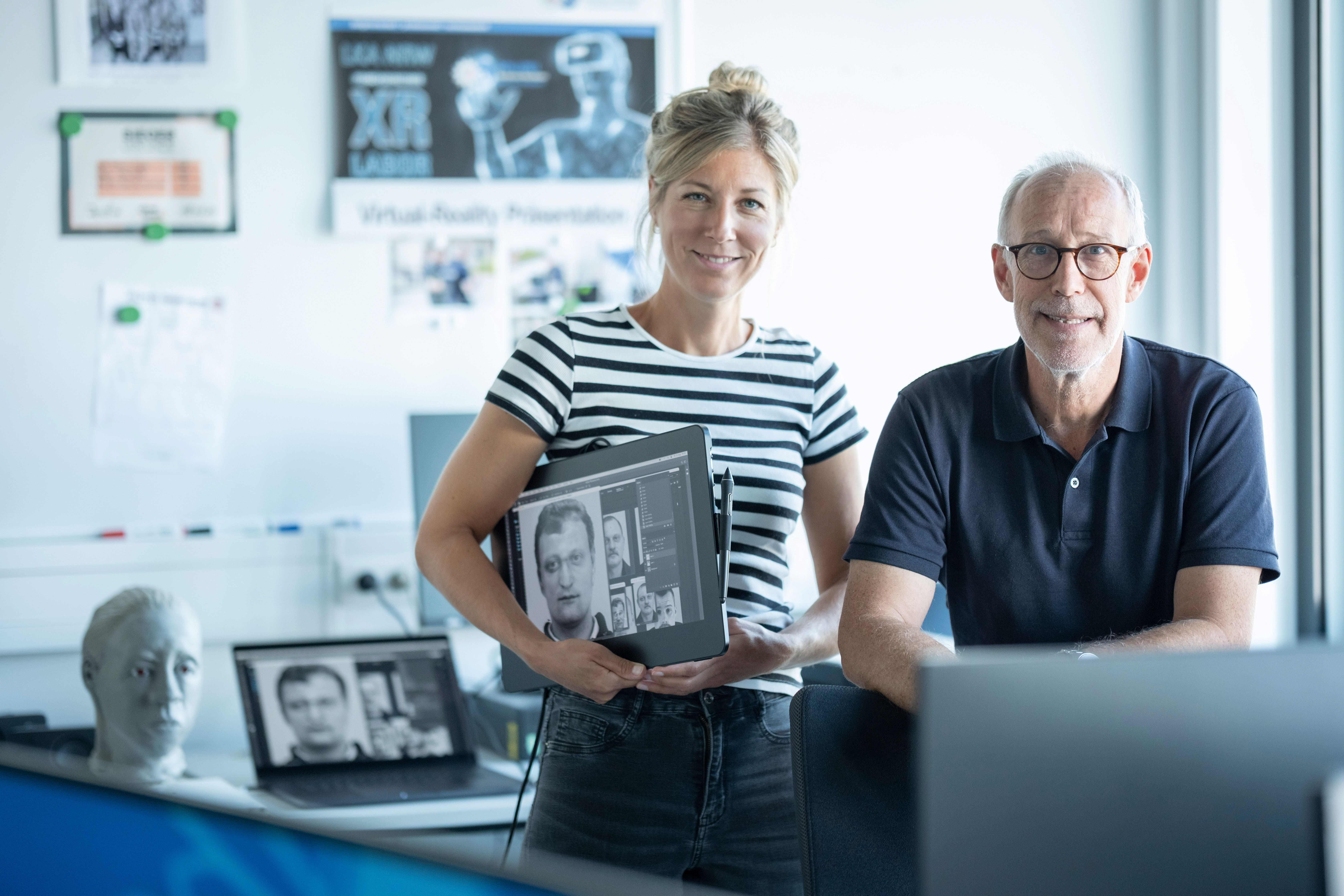 Sketch artist Hanna Mecke and Ingo von Westphal from the LKA NRW are in an office. Hanna Mecke is holding a tablet for sketching sketches under her arm. Both are looking into the camera. Posters can be seen on a white wall in the background.