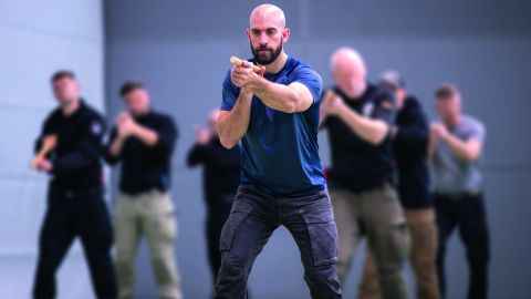 In a training room, a bodyguard can be seen in the foreground holding a baton with both hands in front of him. In the background are other people in the same position.