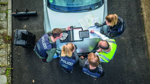 An investigation team from the LKA NRW consisting of two female and three male police officers is standing in front of a silver car. They have spread out equipment and maps on the hood and are looking at them. There is more equipment to the left of the car. The picture was taken from above.