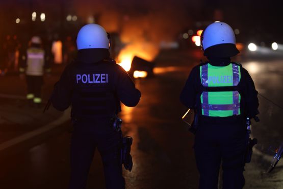 Zwei Polizisten stehen im Dunkeln vor einem brennenden Müllcontainer.